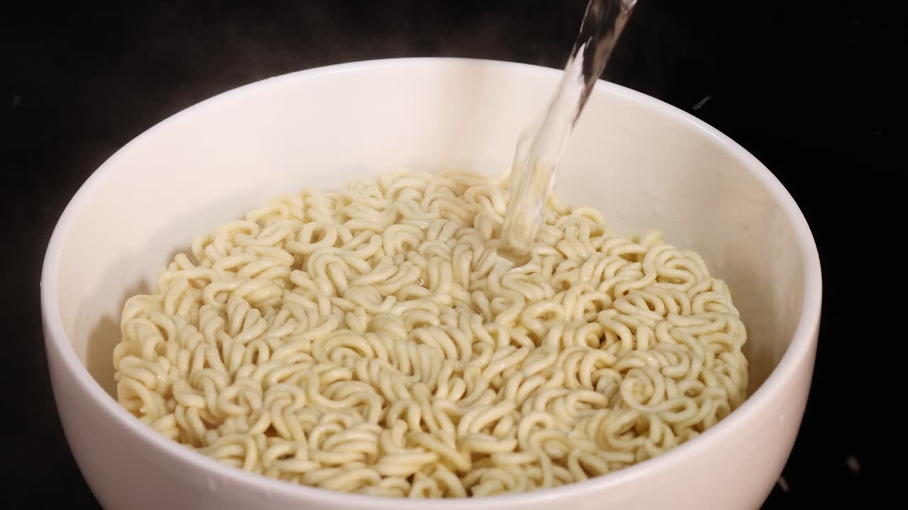A steady stream of hot water is poured onto a block of instant noodles in a white bowl, shot with even lighting and a static overhead camera