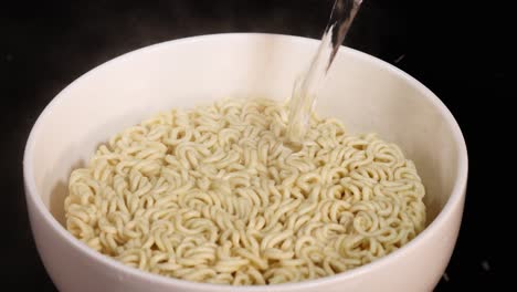a steady stream of hot water is poured onto a block of instant noodles in a white bowl, shot with even lighting and a static overhead camera