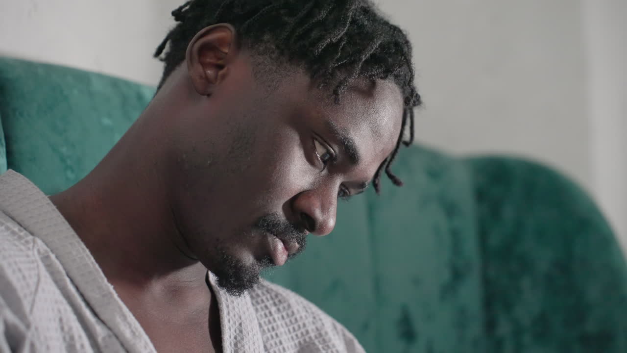 Man in white bathrobe, deep in thought, sitting on bed with green velvet headboard, reading black notebook, focused and relaxed in cozy, soft-lit indoor setting