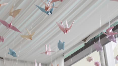 wide shot of colourful pastel origami paper crane birds floating in breeze, hung from roof, blue, yellow, pink and orange