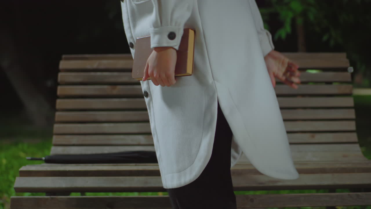 Partial view of girl in white coat holding book while standing beside wooden bench outdoors at night, with blurred background showing greenery, soft bokeh streetlights