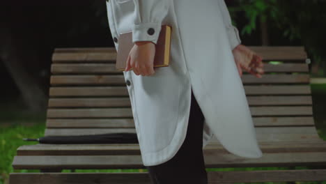partial view of girl in white coat holding book while standing beside wooden bench outdoors at night, with blurred background showing greenery, soft bokeh streetlights