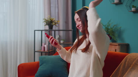 pretty caucasian young woman in headphones listening music dancing, singing in living room at home