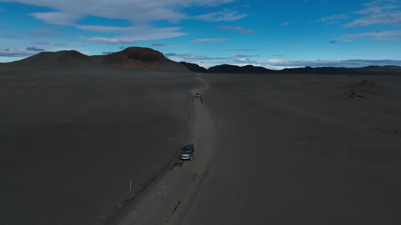 Car drives across Iceland’s vast black-sand highlands toward distant volcanic ridges under shifting blue skies — a remote, cinematic Fjallabak landscape