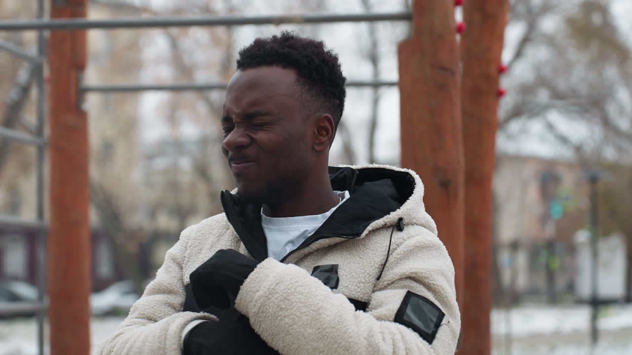 Uomo africano che esegue uno stretching del braccio all'aperto in inverno con il terreno coperto di neve, lo sfondo presenta alberi e neve sfocati, creando un'atmosfera calma per l'allenamento all'aperto.