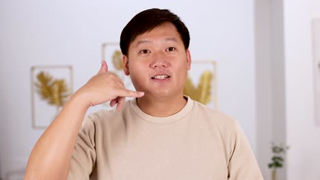 asian man in beige shirt smiles and gestures 'call me' with hand, standing in bright modern room with soft, even lighting and static camera