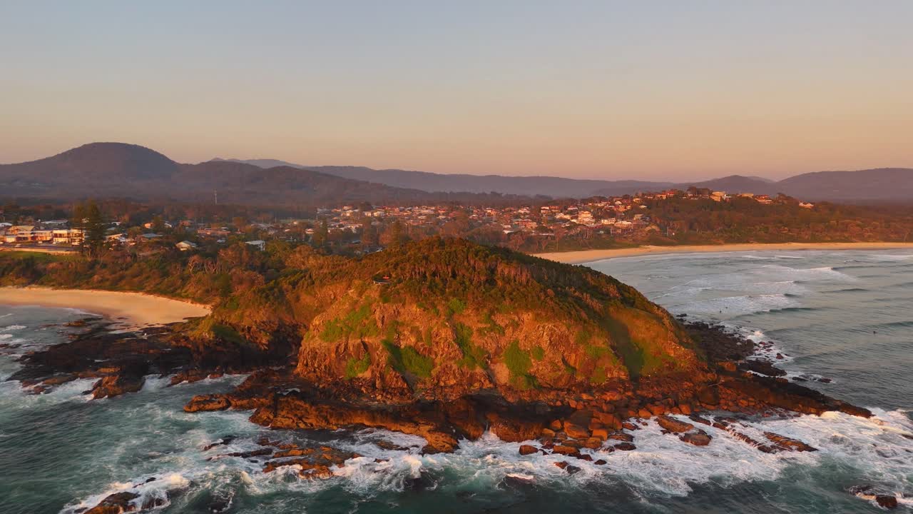 Aerial orbit of rocky peninsula at Scotts Head bathed in golden sunset light over ocean