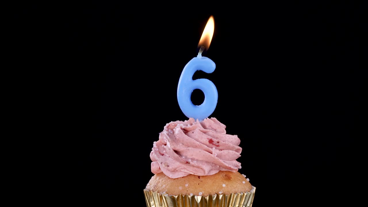 A single cupcake with pink frosting and a lit blue number six candle is shown against a black background, illuminated by the candle flame in a static shot