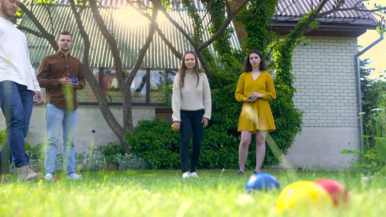 кавказская молодая женщина бросает желтый шар для петанка в парке в солнечный день, пока ее друзья ждут своей очереди