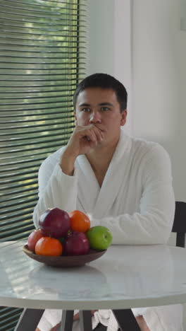 hombre se sienta pensando en la mesa de comedor. tipo en túnica de terry piensa en la nutrición saludable esperando el desayuno. variedad colorida de frutas frescas gracias mesa