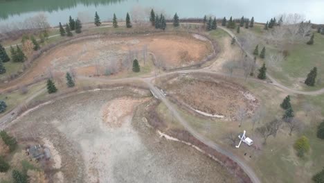 aerial-rise-dolly-roll-fly-over-birds-eye-view-dry-ponds-with-tree-trimming-pruning-man-lift-articulated-arm-platform-on-a-fall-afternoon-scattered-pine-trees-bare-varying-trees-empty-path-by-river3-3