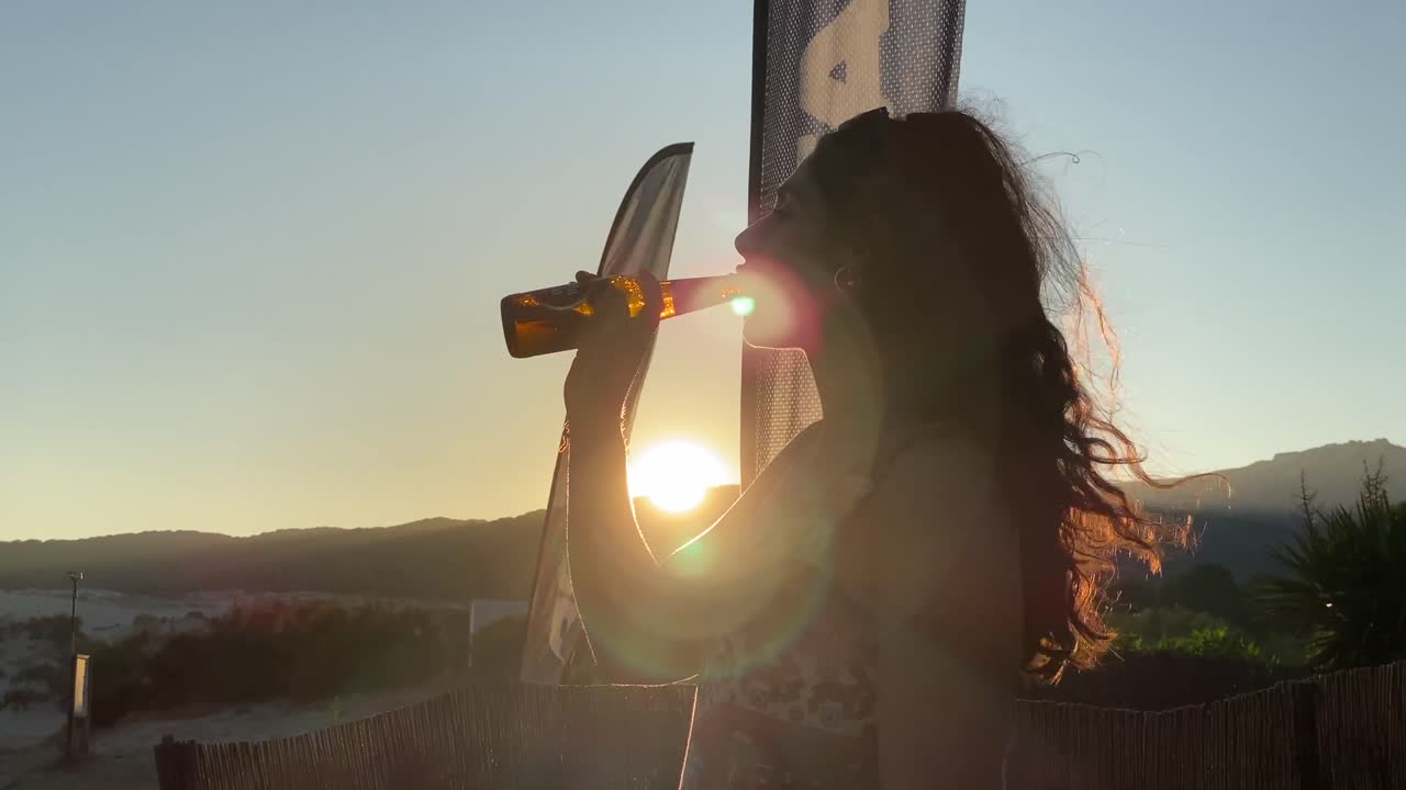 Giovane donna che beve birra con il tramonto alle spalle in spiaggia.
