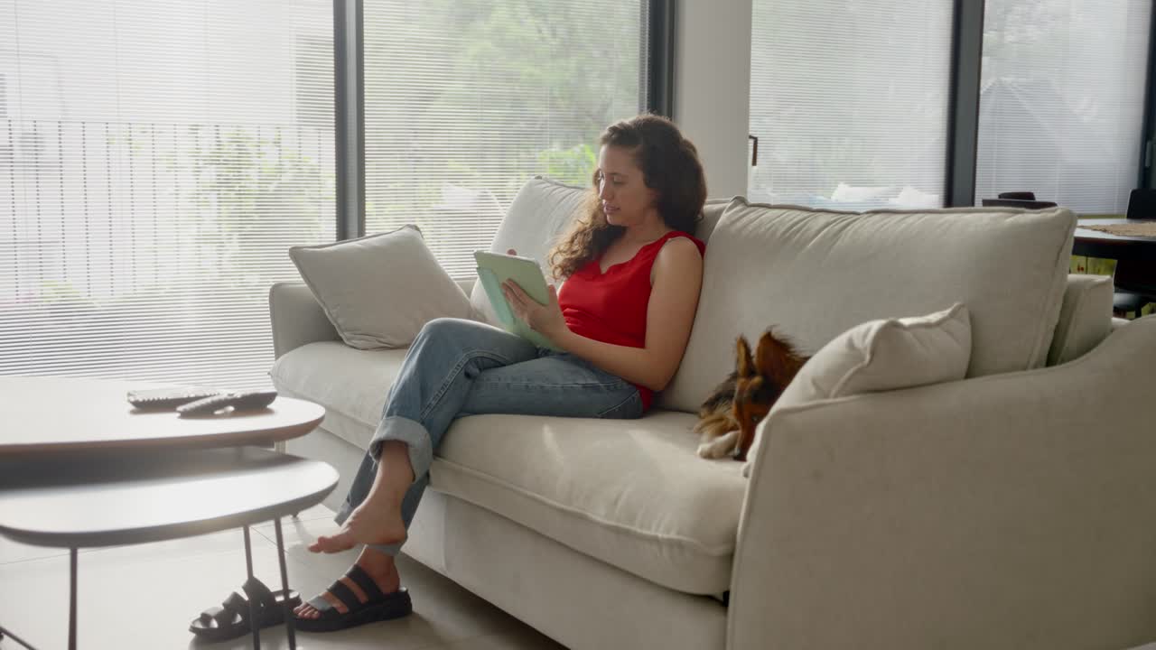 Young woman sitting on couch in apartment with her dog writing on tablet with stylus pen. Drawing or designing in creative form or occupation. Digital art. Working from home happy and cheerful