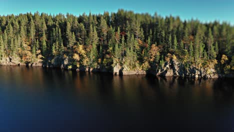 a dense forest with mixed autumn foliage stands tall along the edge of a deep, dark lake with rocky shores