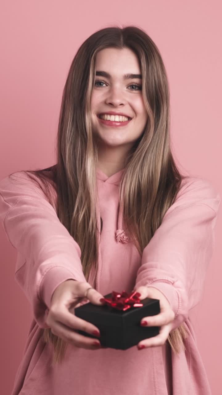 Cheerful young woman giving gift box in pink studio