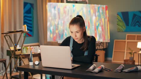 una morena sonriente se sienta en un escritorio frente a una computadora portátil en un estudio de arte y muestra pinturas para la venta, alrededor yace un pincel, pinturas de acuarela en un tubo, en el fondo una pintura multicolor