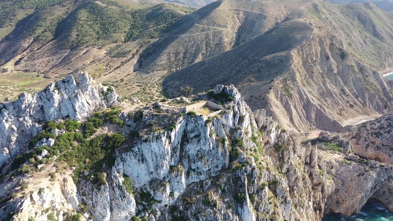 Vista aerea cinematografica con drone di un'enorme scogliera calcarea che si affaccia su montagne costiere a strati e acque turchesi del Mediterraneo nel Marocco settentrionale