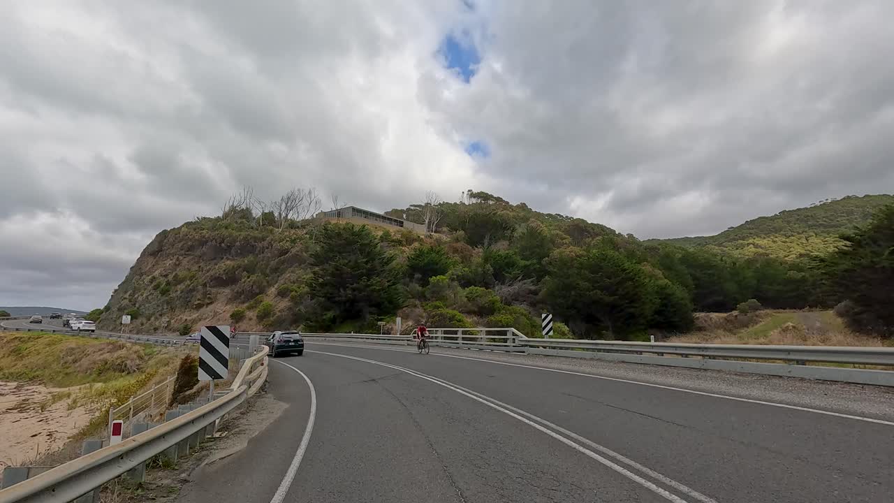 A cyclist and multiple vehicles travel along a winding coastal highway under overcast skies, with lush hills and ocean views on the Great Ocean Road