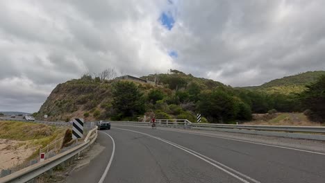 a cyclist and multiple vehicles travel along a winding coastal highway under overcast skies, with lush hills and ocean views on the great ocean road