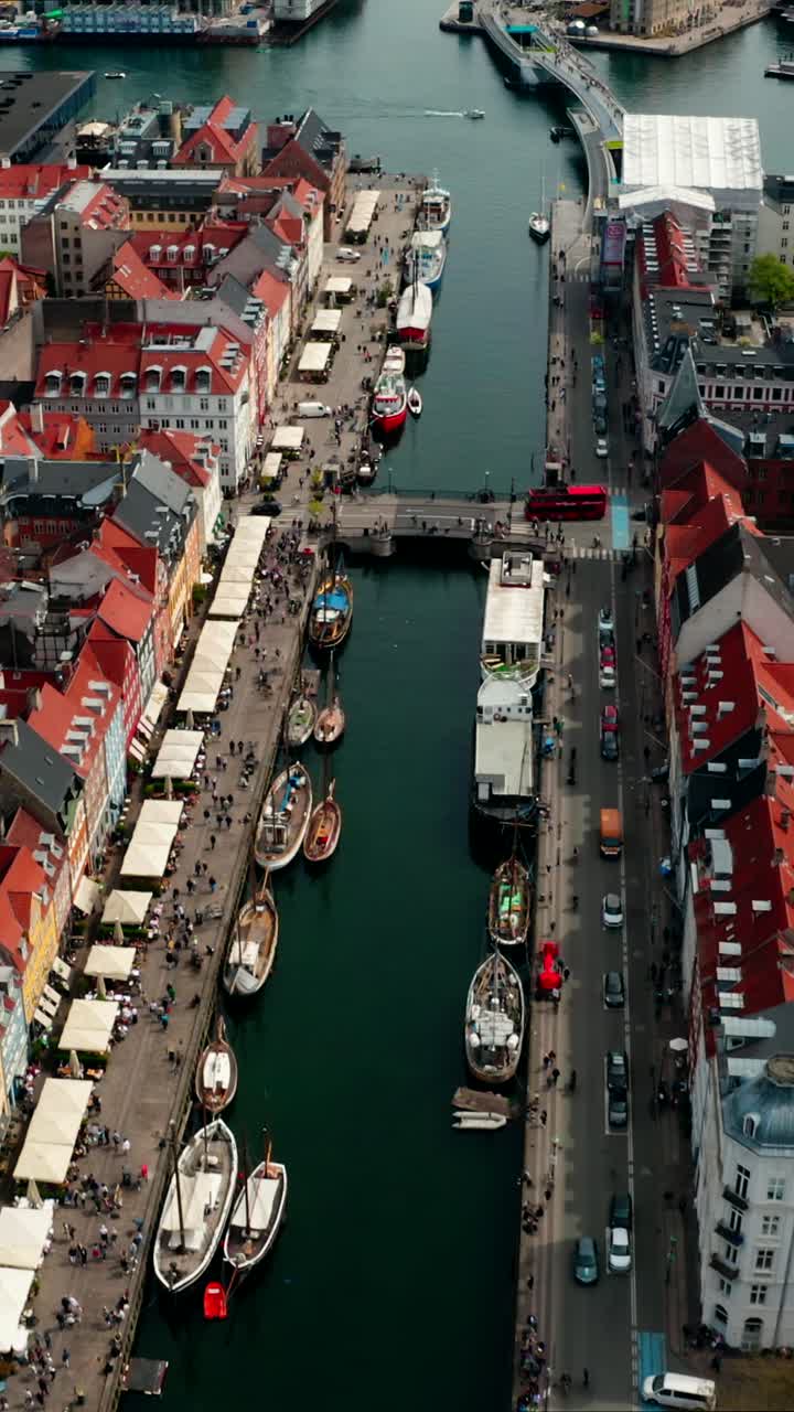 Vertical high view of Nyhavn street, Copenhagen. With brightly coloured townhouses, a canal with historic boats, and tourists enjoying the sunny day out on the waterfront.