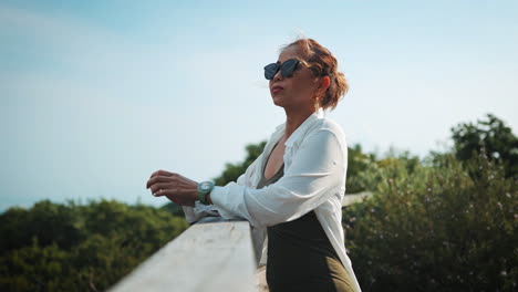 woman stands by a railing, looking at the lush green landscape and blue sky