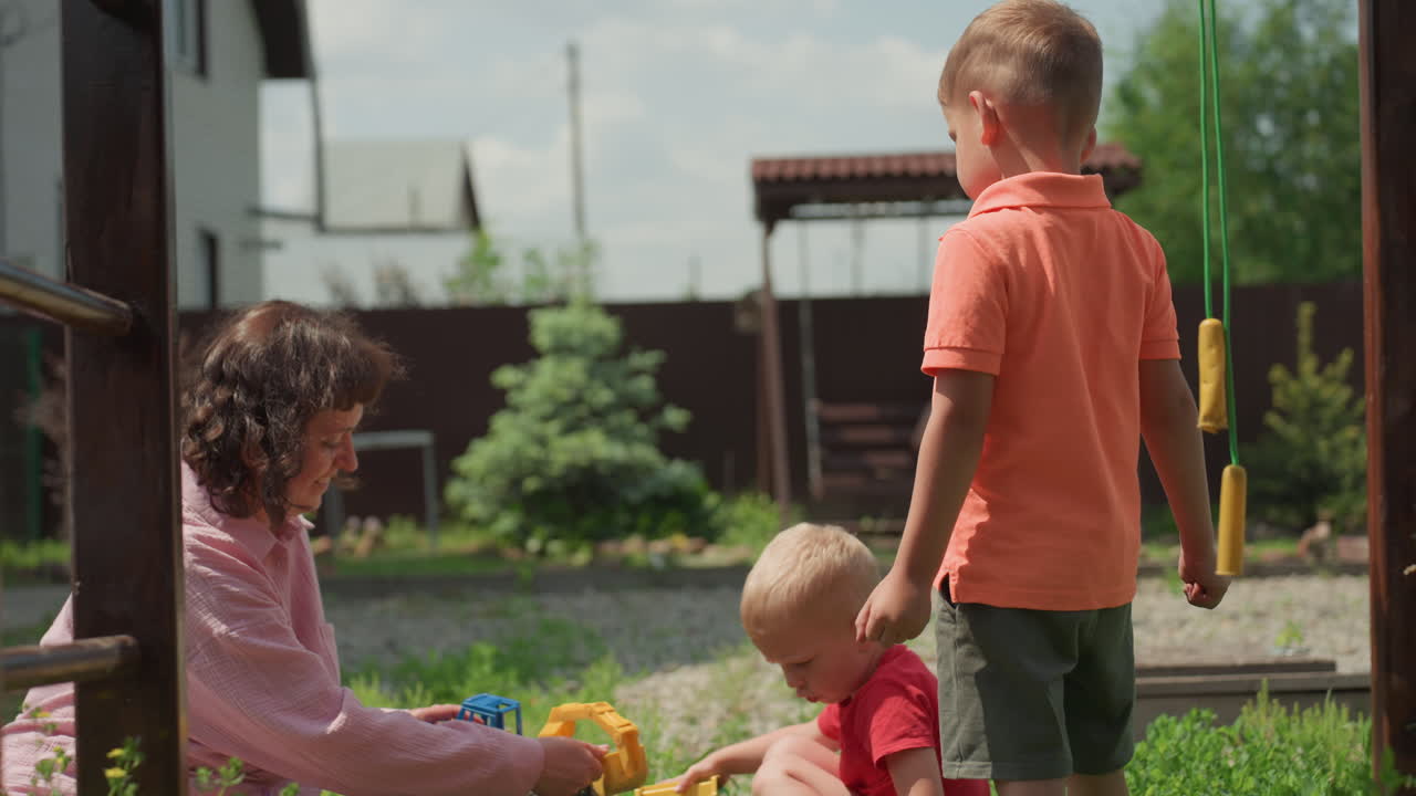 Child Receives Toy Amid Family Outdoor Setting, Parent Gently Gives Toy As Brother Watches Attentively Outside, Tender Moment Of Mother Offering Toy To Young Child With Older Sibling Beside Them