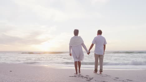 Back-view-of-hispanic-just-married-senior-couple-walking-on-beach-at-sunset