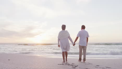 back view of hispanic just married senior couple walking on beach at sunset