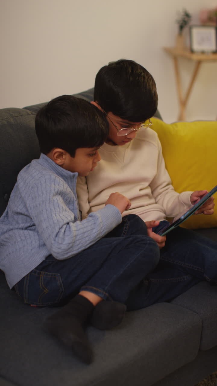 Video verticale di due giovani ragazzi seduti sul divano a casa che giocano o guardano contenuti in streaming insieme su un tablet digitale 2
