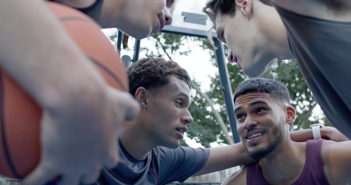 Diverse male team in tanks huddling with left player gripping basketball nodding preparing at court