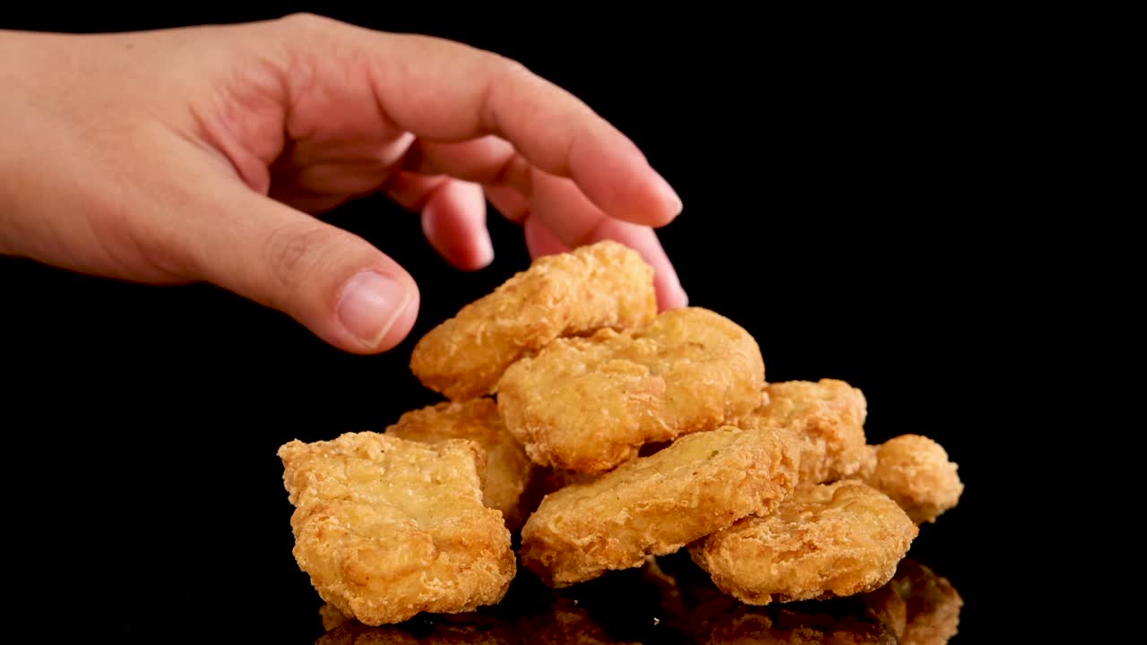 A hand reaches in to pick up a spicy, crumbed chicken nugget from a stacked pile on a reflective black surface, under bright studio lighting