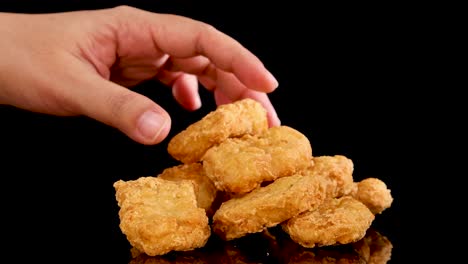 a hand reaches in to pick up a spicy, crumbed chicken nugget from a stacked pile on a reflective black surface, under bright studio lighting