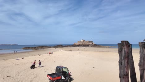 wooden pillars on the beach with seagull perched on top, distant fort national, and walled city with people, saint malo, brittany, france