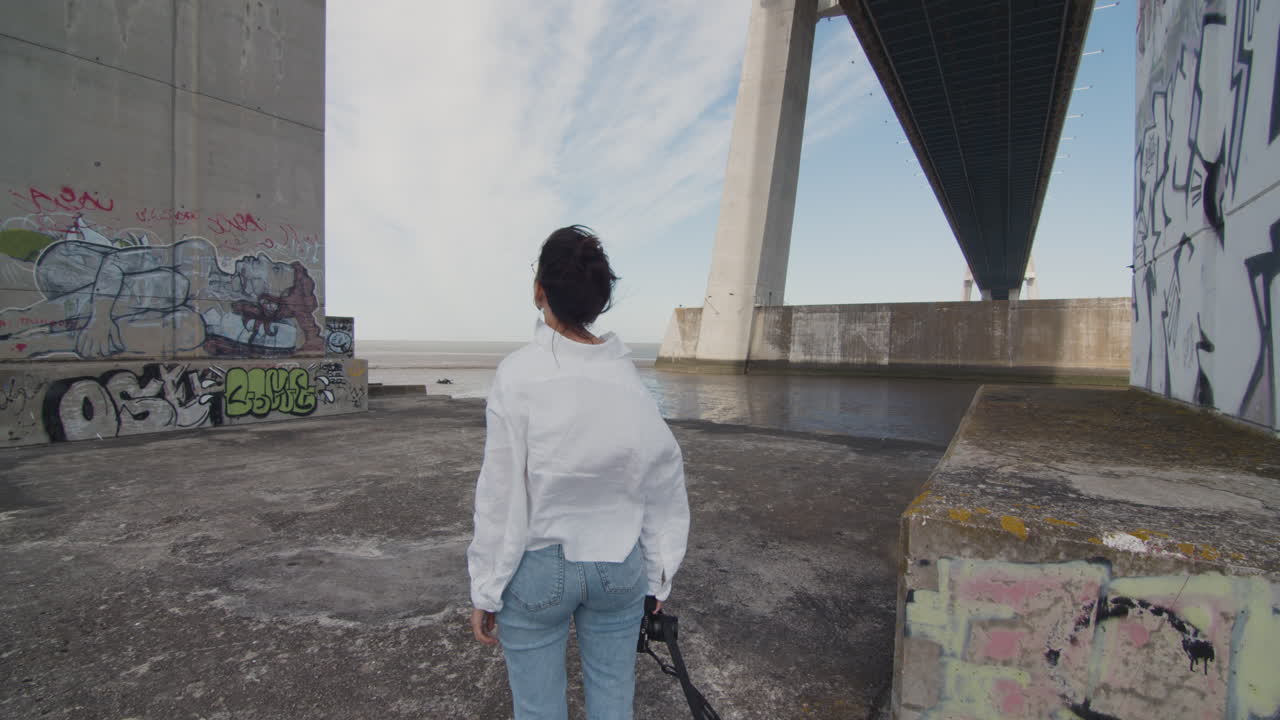 Medellång bild av en ung kvinna som betraktar det urbana, graffitifyllda landskapet. Starka vindar under Vasco da Gama-bron. Urban isolering. Förundran och vördnad i ensamhet.