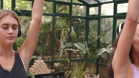 practicing yoga, woman stretching arms up in glass house with lush plants, copy space