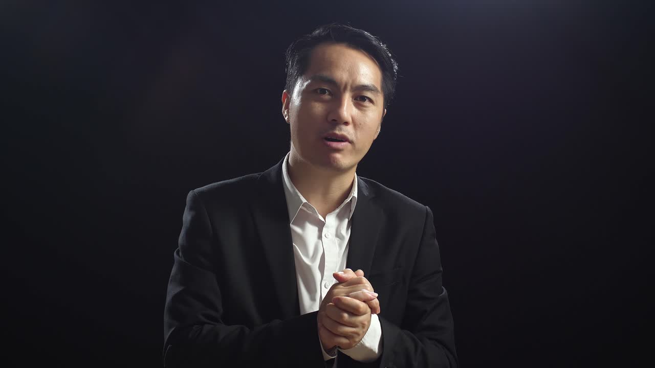 Close Up Of Asian Speaker Man In Business Suit Holding His Hands Together And Showing Index Fingers Up While Speaking In The Black Screen Studio