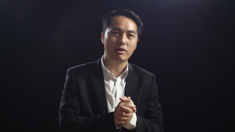블랙 스크린 스튜디오에서 말하는 동안 손을 함께 잡고 지표손가락을 보여주는 비즈니스 슈트를 입은 아시아 스피커의 클로즈업