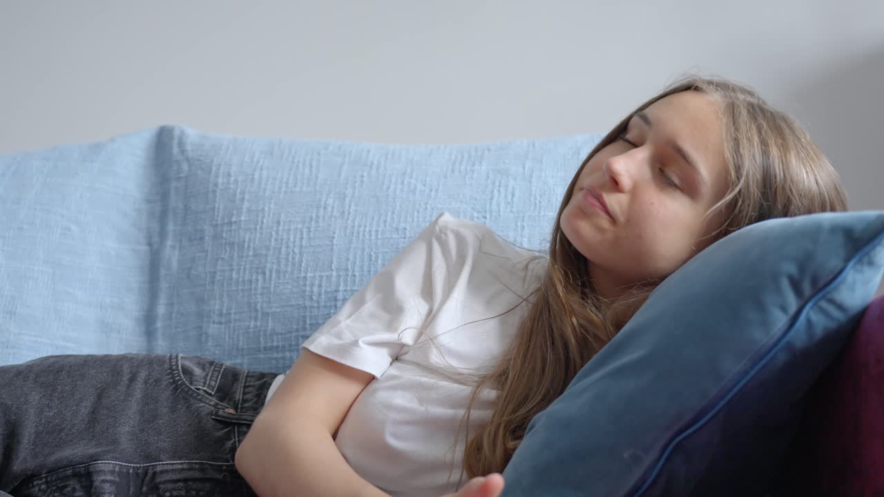 Tired girl resting on sofa at home, pointing remote at tv to change channel