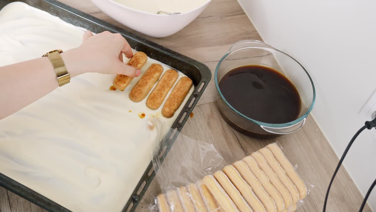 Top-down shot of unrecognizable person assembling tiramisu by layering coffee-soaked ladyfingers over mascarpone cream, bowl of espresso and biscuits nearby, real time, kitchen prep scene