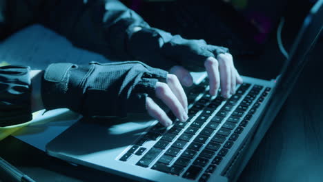 closeup shot of human hands in fingerless gloves typing on grey keyboard in dark room while hacking or coding at night