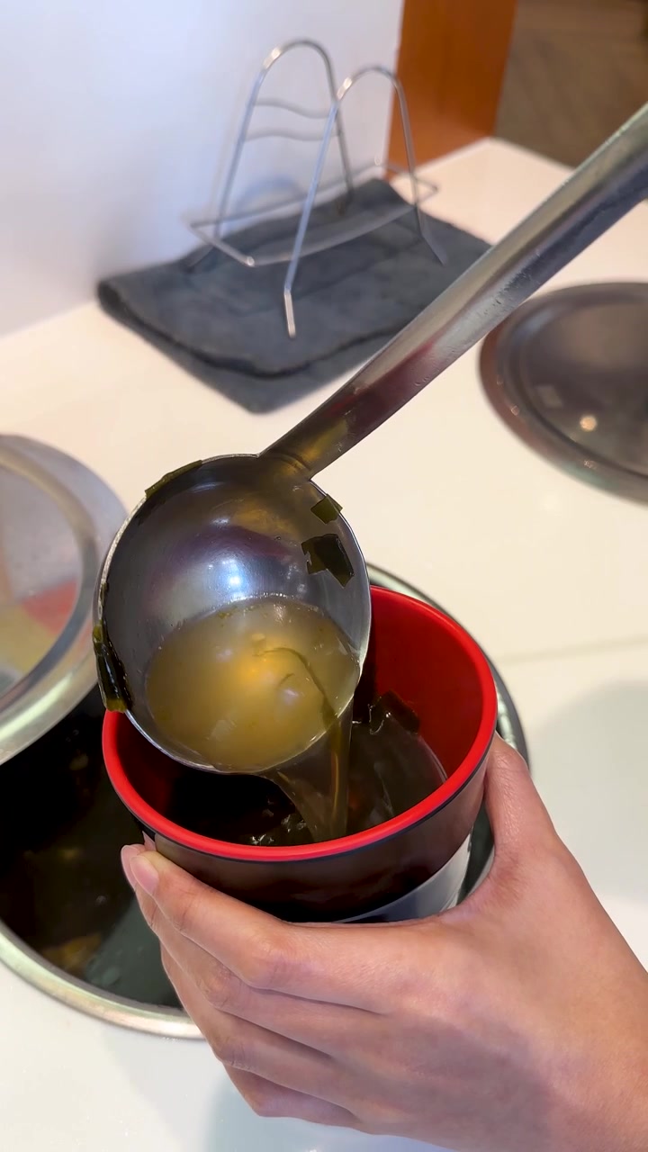 Hand ladles steaming seaweed soup into red bowl, bright indoor kitchen lighting, close-up view