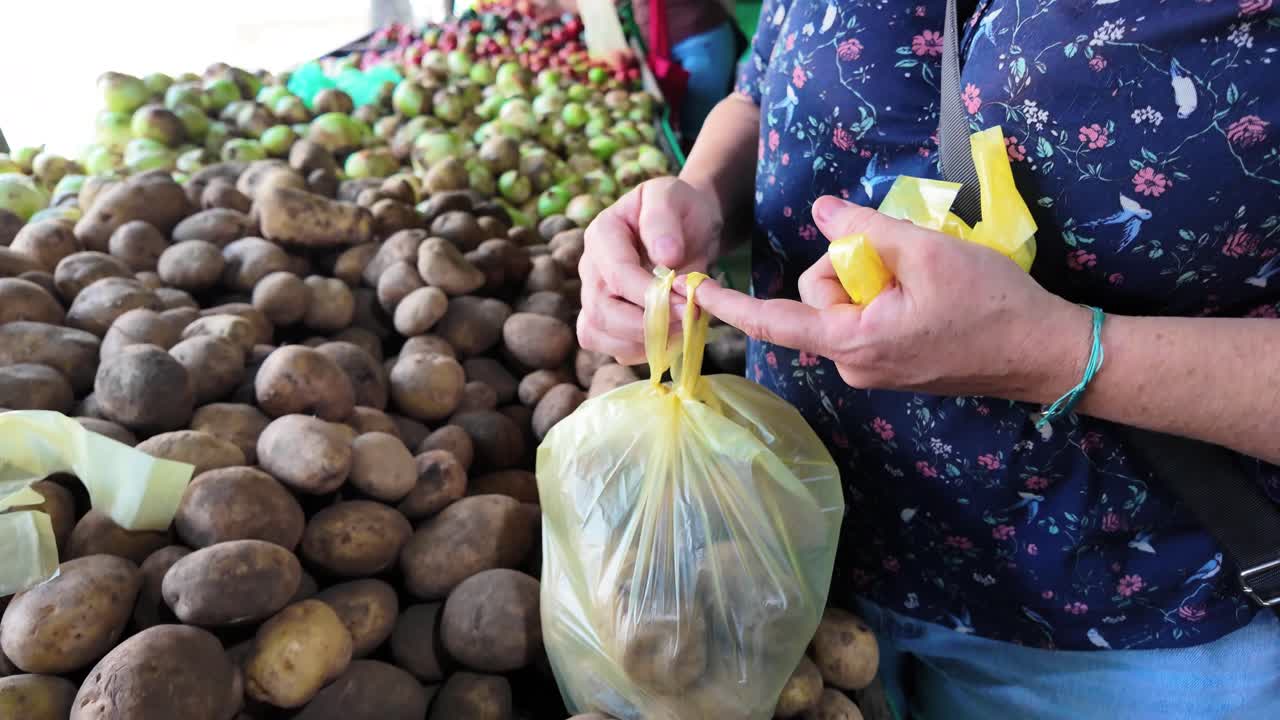 A woman picks up a bag of potatoes to weigh and buy it at the local food market.