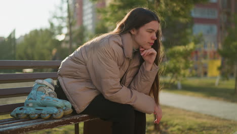 una joven con una chaqueta de melocotón, camisa interior gris y pantalones negros se sienta en un banco del parque, descansando su barbilla en una mano en profundo pensamiento. sus patines están colocados a su lado