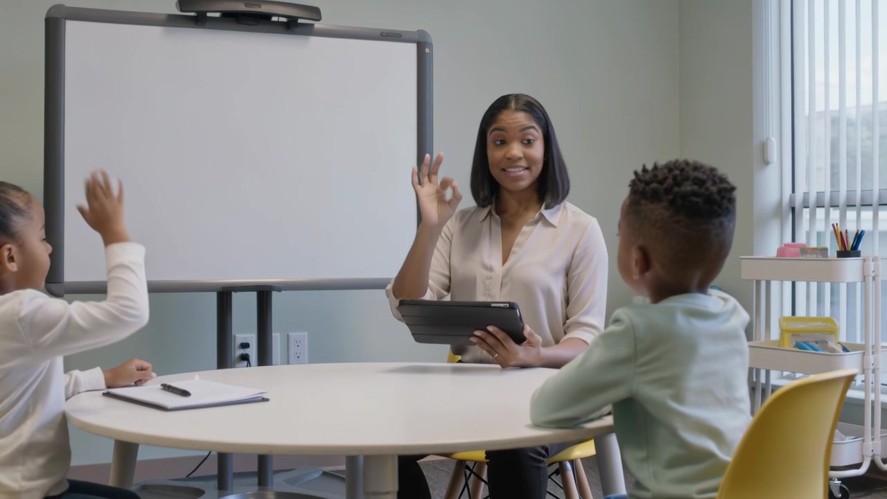 Gesturing toward whiteboard tutor guiding kids around table, managing turns with tablet in blouse