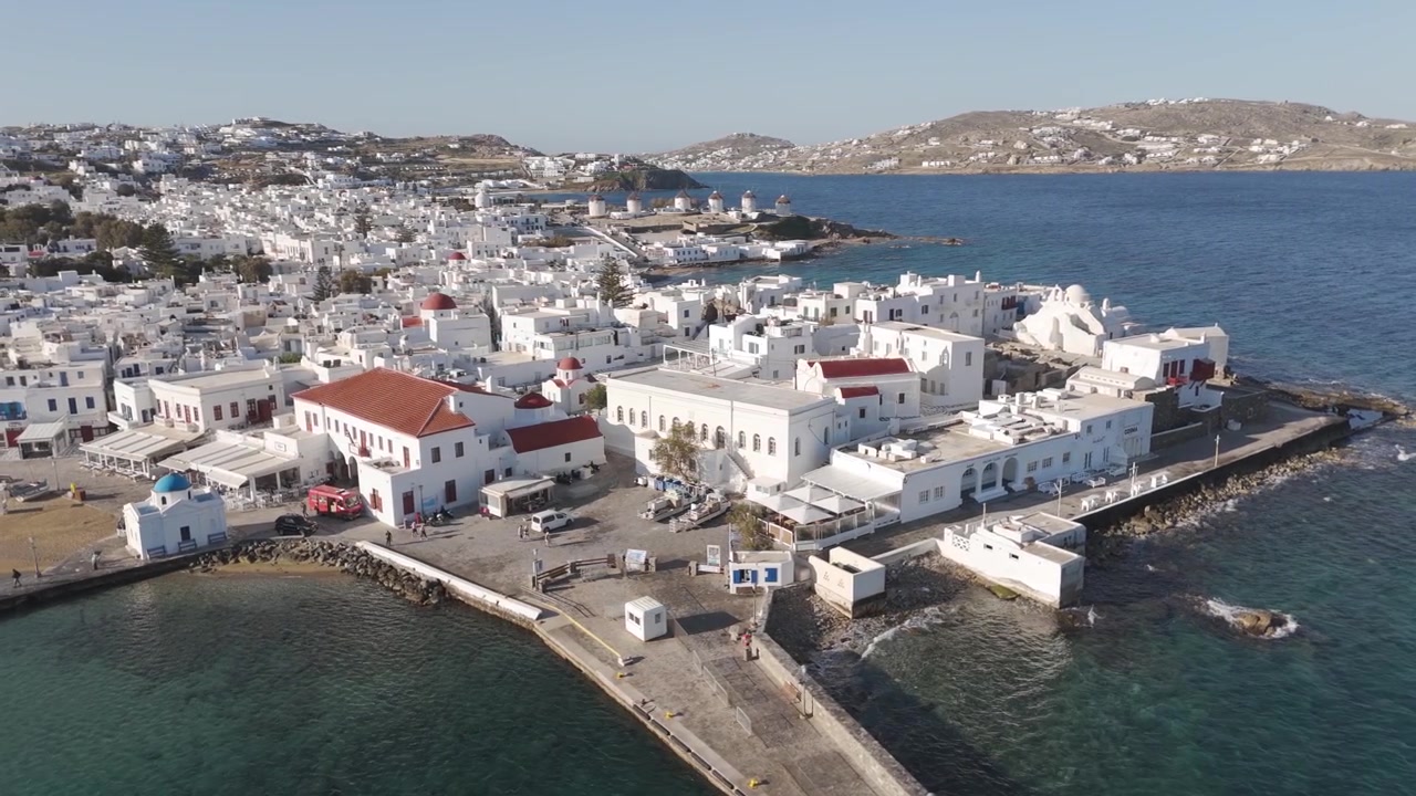 Veduta aerea soleggiata del porto della città di Mykonos con i suoi iconici edifici bianchi e il Mar Egeo. Dolly circolare.