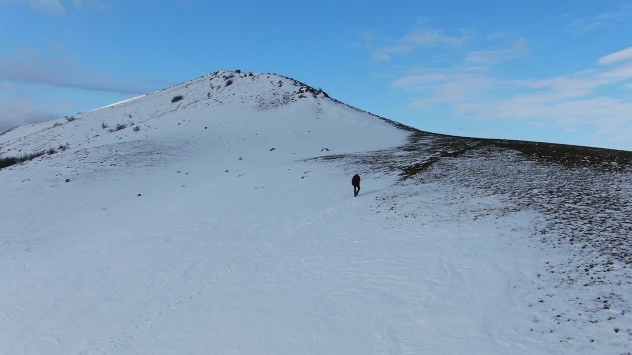 Aerial view as a Man with a backpack walks along the slope of a hill that is covered with snow