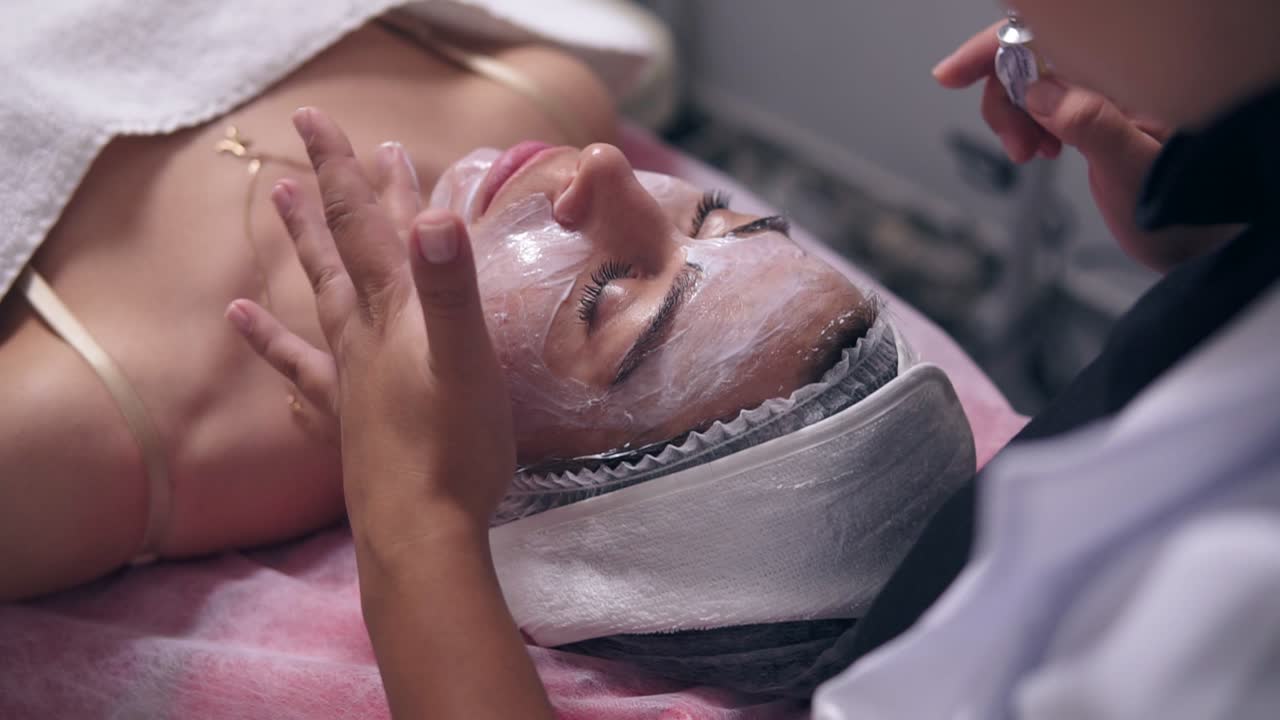 Close Up view of young woman lying on the couch while professional cosmetologist is applying special treatment on woman's face