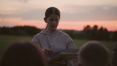tutor holding binder interacting with seated group as insect buzzes around colleague passing by adjusting glasses capturing candid outdoor lesson moment under soft sunset light