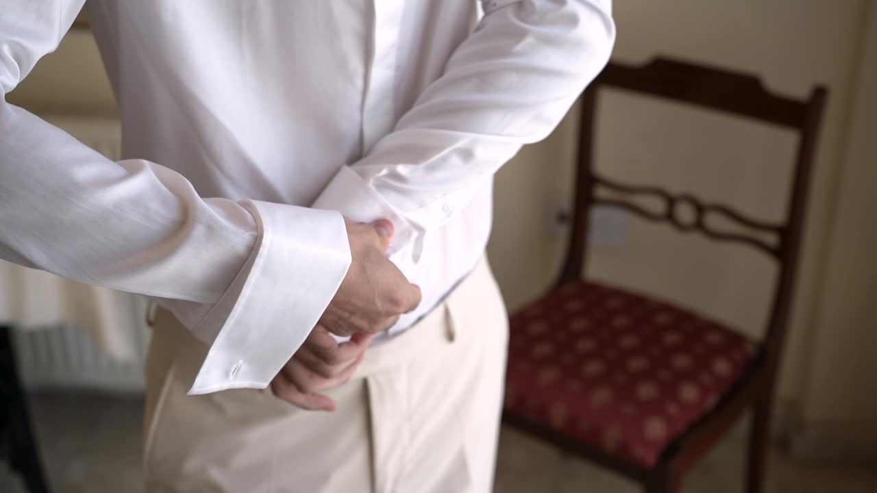 Uno sposo si sistema i polsini della camicia in una stanza. Si prepara per il giorno del suo matrimonio, concentrandosi sull'indossare abiti eleganti. L'emozione cresce con l'avvicinarsi dell'ora della cerimonia.