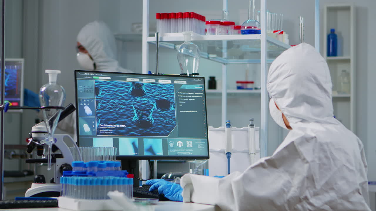 ppe 의복을 입은 화학자가 컴퓨터에서 타이핑하고 바이러스 개발을 확인합니다.